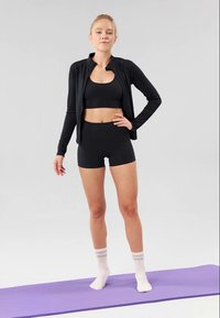 Svart idrottsjacka med dragkedja i kragen, matchande crop top och shorts, bärs med vita strumpor på en lila yogamatta.