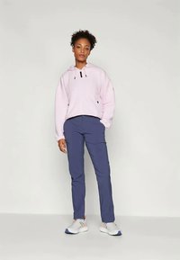 Felpa rosa chiaro con finitura testurizzata, dettagli neri e lacci regolabili, abbinata a pantaloni blu navy e sneakers bianche.