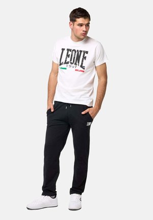 Joven con camiseta blanca Leone 1947, pantalones deportivos negros con logo, zapatillas blancas, de pie con las manos en los bolsillos, mirando hacia un lado.
