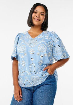 Blusa ricamata azzurro chiaro con maniche corte a sbuffo, caratterizzata da motivi floreali e circolari tagliati con dettagli in pizzo, abbinata a jeans di denim.