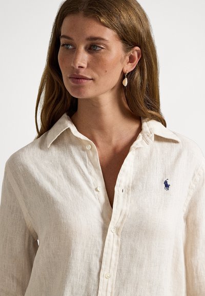 Linen button-up shirt in light beige with a textured finish and a small blue logo on the chest. Classic collar and white buttons.