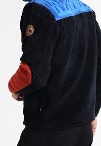 Pullover in tessuto felpato con colletto blu, corpo nero e maniche rosse. Presenta spalle testurizzate e logo ricamato sul braccio sinistro. Tessuto morbido e caldo.