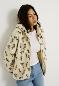 Veste en polaire à motif floral crème, avec fermeture éclair, capuche et deux poches avant. Tissu texturé avec des accents roses et verts.