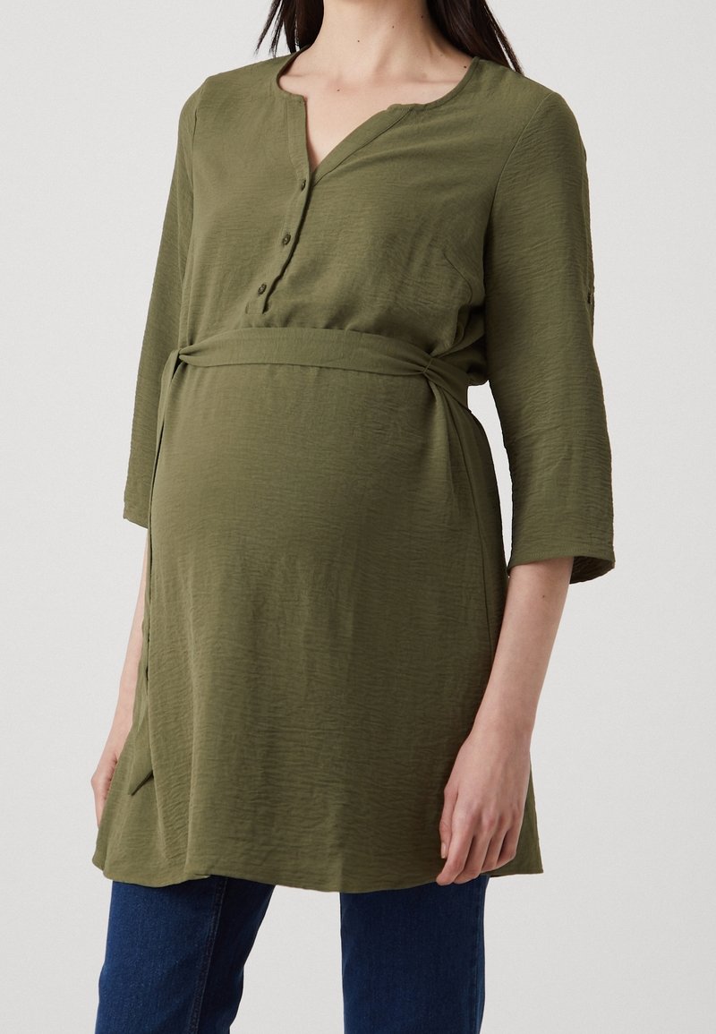 Femme enceinte portant une tunique de maternité vert olive à manches trois quarts et un jean bleu, debout devant un fond clair uni.