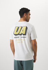 Hvid bomulds t-shirt med stort sort og gult "UA BODY SHOP" logo på ryggen med teksten "Kvalitetstjeneste siden '96" nedenfor.