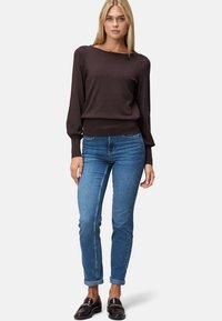 Femme blonde debout, portant un pull à manches longues marron foncé, un jean bleu retroussé et des mocassins noirs avec des accents dorés.