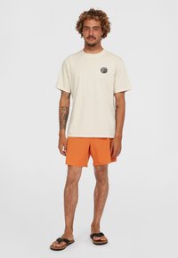 Camiseta de algodón color beige claro con mangas cortas y un logo redondo en el pecho, combinada con unos shorts de color naranja vibrante y chanclas negras.