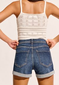 Shorts de mezclilla con un lavado azul, puños enrollados y dos bolsillos traseros. Combinados con un top corto de crochet blanco que presenta patrones intrincados.