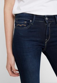 Jeans de mezclilla azul oscuro con corte slim, que presentan costuras en contraste, un bolsillo frontal con emblemas decorativos y un cierre clásico de botón.
