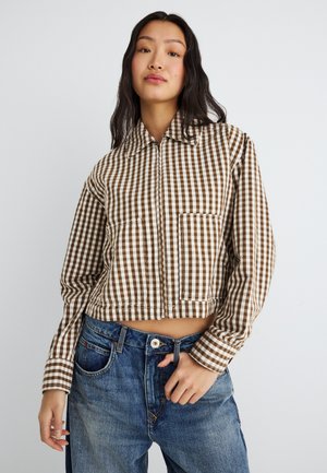 Γυναίκα με μακριά σκούρα μαλλιά, ντυμένη με ένα κοντό καφέ και άσπρο καρό τζάκετ και ψηλόμεσα ξεθωριασμένα μπλε τζιν, σε απλό φόντο.