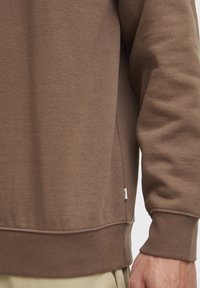 Sudadera marrón con una textura suave, puños acanalados y una etiqueta en la costura lateral. Presenta un corte relajado y un diseño simple.