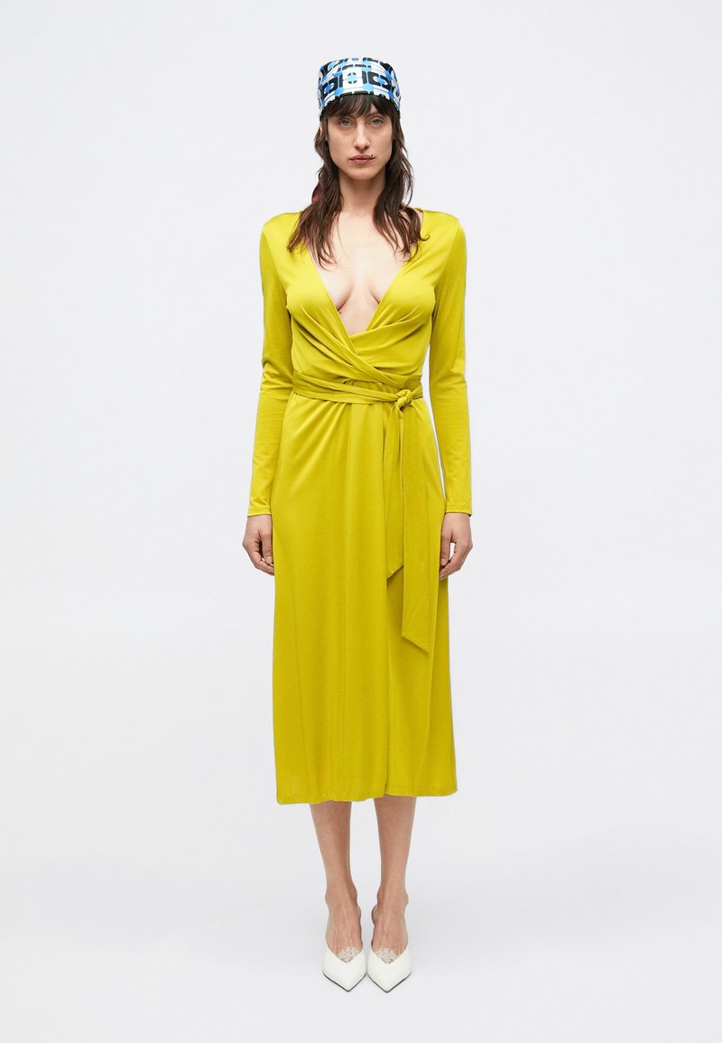 Rochie galbenă tip wrap cu mâneci lungi și decolteu adânc în V, cu talie legată și textură fină. Asortată cu pantofi cu toc alb, cu vârf ascuțit, și un batic cu model.