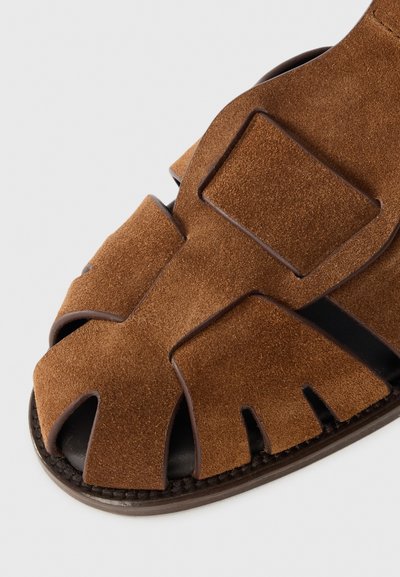 Brown suede sandal with open-toe design, featuring cut-out accents and dark leather trim around the sole for added contrast and detail.
