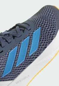 Zapatilla de running con parte superior de malla gris, acentos sintéticos azules, cordones planos y una entresuela blanca con una suela amarilla.