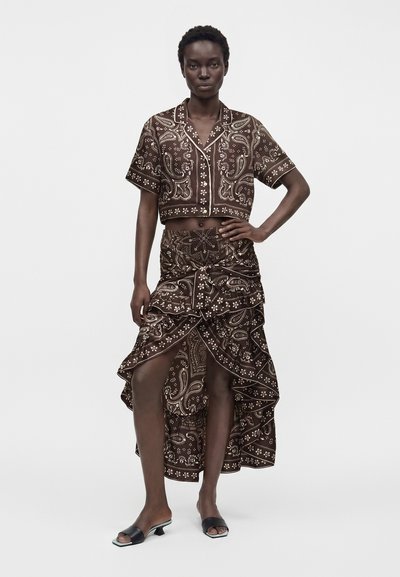 Femme portant une chemise courte marron à manches courtes à motifs et une jupe asymétrique assortie avec des volants, debout, une main sur la hanche.