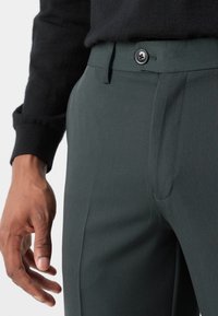 Tmavě zelené kalhoty z hladkého materiálu, s knoflíkovým zapínáním, přiléhavým střihem a viditelnými bočními kapsami. Ruka spočívá na látce.