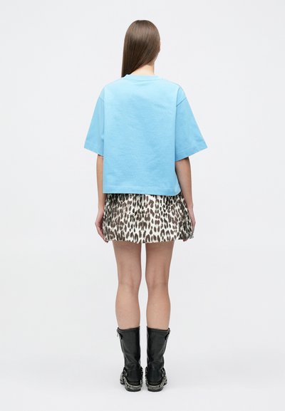 Γυναίκα που κοιτάζει προς τα πίσω, ντυμένη με ανοιχτό μπλε κοντομάνικο πουκάμισο, φούστα με λεοπάρ μοτίβο και μαύρα μποτάκια μέχρι τη μέση της γάμπας με λεπτομέρειες από αλυσίδα.