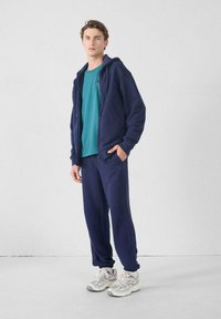 Marineblauwe hoodie met rits en zijzakken, gecombineerd met een teal t-shirt en bijpassende marineblauwe joggingsbroek. Grijze sportschoenen maken de outfit compleet.