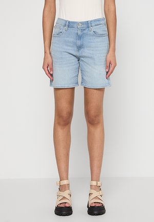 Short en denim bleu clair porté par une personne debout, associé à des sandales beiges épaisses avec des brides à la cheville sur un fond blanc uni.