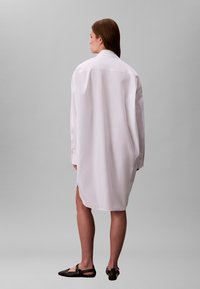 Fehér, túlméretes ing klasszikus gallérral, hosszú ujjakkal és oldalsó sliccekkel. A sima anyag a combközépig ér, laza szabásra tervezve.