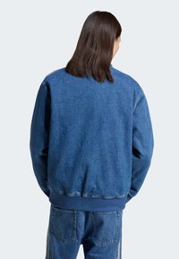 Chaqueta de mezclilla en azul medio con un corte relajado. Presenta puños y dobladillo acanalados. La vista trasera muestra una textura suave sin detalles adicionales.