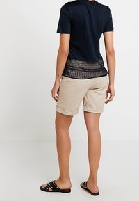 Osoba stojí zády k fotoaparátu, na sobě má černé tričko, béžové šortky a černé pantofle se hroty, což ukazuje na ležérní outfit.