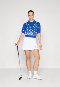 Blå strikket polo skjorte med hvite blomster mønster, sammen med en hvit golfskjørt og hvite joggesko. Golfkølle i modellens hånd.