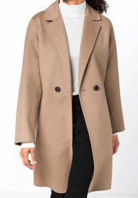 Béžový kabát z vlněné směsi s dvouřadým zapínáním, s klopovým límcem, černými knoflíky a dlouhými rukávy; nošený přes bílý rolák.