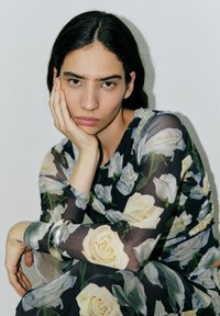 Kleid mit einem schwarzen, transparenten Blumenmuster, das große weiße Rosen zeigt, langen Ärmeln und einer figurbetonten Silhouette. Silberne Armreifen am Handgelenk.