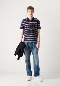 Stripet navy polo skjorte, kortermet, laget av bomull. Kombinert med lyseblå jeans og hvite sneakers. Svart hettegenser lagt over armen.