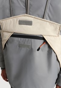 Grå vadderad jacka med en beige klaff med kardborre, en zip-fick på framsidan och en orange drarem for enkel åtkomst.