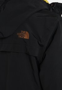 Czarna wodoodporna kurtka z żółtym wnętrzem, wysokim kołnierzem i brązowym logo na górnej części pleców. Teksturowana tkanina i minimalistyczny design.