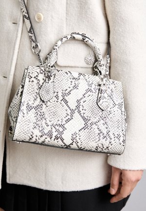 Vrouw houdt een witte en grijze Michael Kors handtas met slangenhuidpatroon vast, met dubbele handvatten en een schouderriem, draagt een crèmekleurige jas.