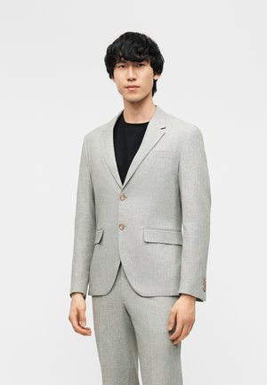 Νέος άνδρας που φοράει ανοιχτογριζό δίκουμπο κοστούμι με τσέπες με καπάκια πάνω από μαύρο μπλουζάκι με στρογγυλή λαιμόκοψη, στέκεται μπροστά σε απλό φόντο.