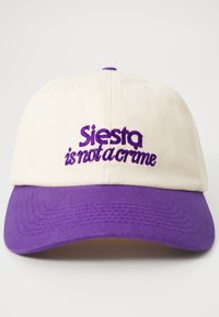 Béžová baseballová čiapka z bavlnenej látky s fialovým okrajom. Obsahuje vyšívaný nápis "Siesta nie je zločin" vo fialovej farbe.