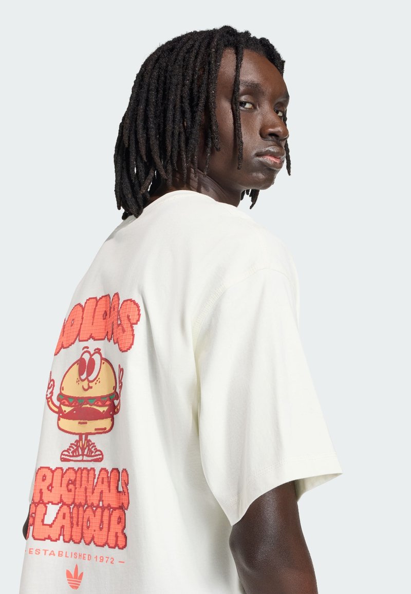 Mann mit mittellangen Dreadlocks, der ein weißes T-Shirt mit einem Cartoon-Burger und dem Text "Originals Flavour" auf der Rückseite trägt.