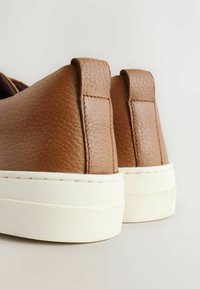 Zapatillas de cuero marrón con acabado texturizado, que presentan una suela de goma blanca y una pestaña en el talón para facilitara ponerlas, centrado en la vista trasera.