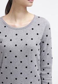Femme portant un pull gris à motif de pois noirs et à encolure ronde, visible des épaules jusqu'au haut de la poitrine.