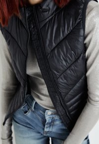 Gilet noir rembourré avec un motif matelassé, col haut et fermeture éclair, porté sur une chemise à manches longues claire et un jean bleu.