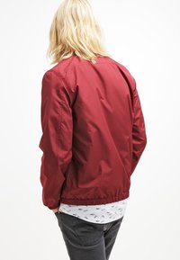 Giacca bomber rossa con polsini e orlo elasticizzati, realizzata in un materiale liscio e leggero. Indossata sopra una camicia bianca a fantasia.