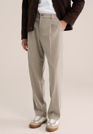 Homme portant un pantalon plissé beige, des baskets blanches avec des accents beiges, une chemise blanche et une veste marron foncé avec une main dans la poche.