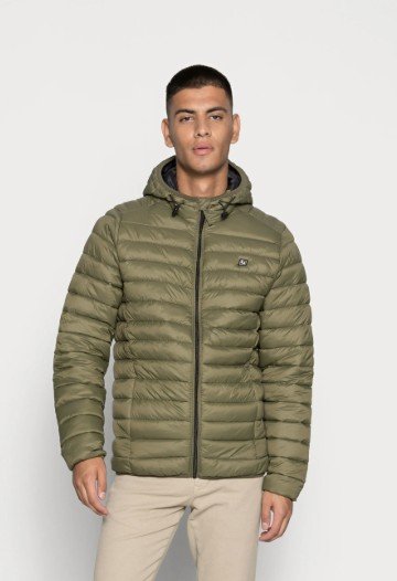Homme portant une veste matelassée à capuche olive avec une fermeture éclair devant et un pantalon de couleur claire, debout devant un fond uni.