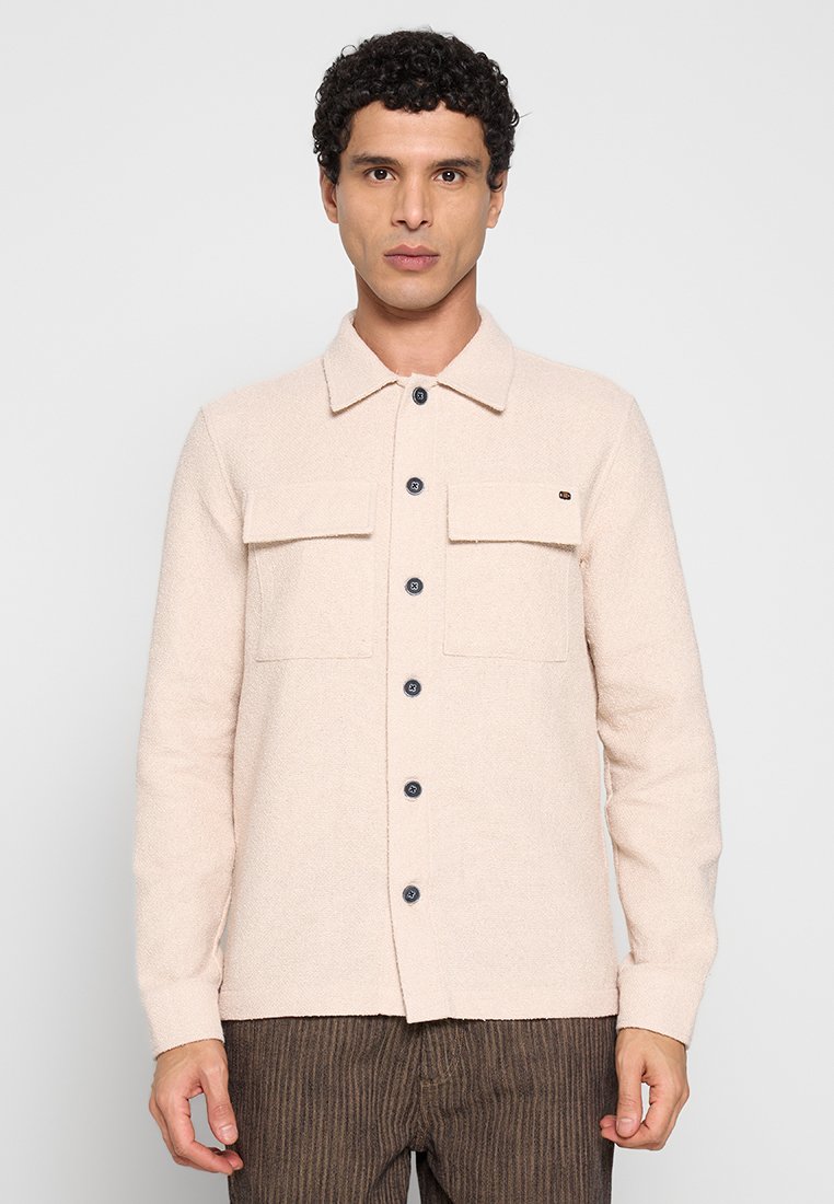 Lichtbeige blouse met knopen en een gestructureerde stof, voorzien van twee borstzakken en donkere knopen. Lange mouwen en een gestructureerde kraag.