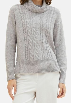 Kvinde iført en lysegrå kabelstrikket rullekravesweater kombineret med off-white bukser, vist fra midt på brystet til hofterne.