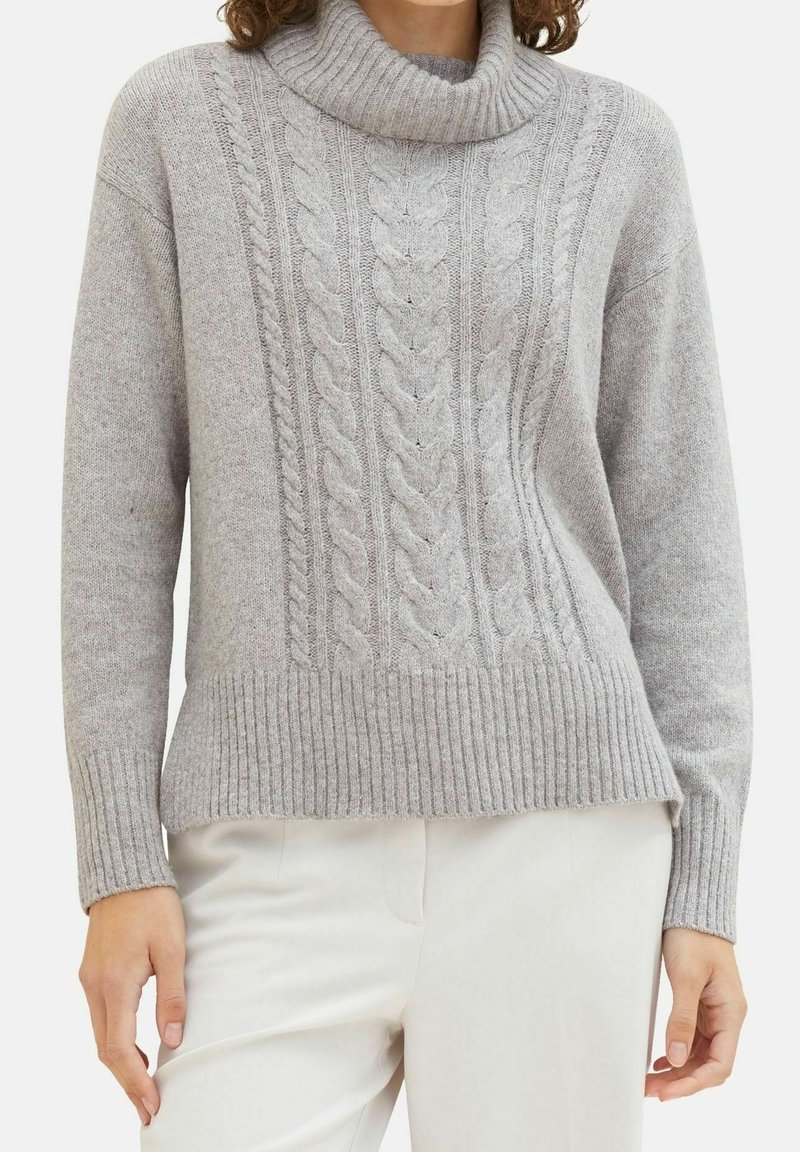 Femme portant un pull à col roulé en maille torsadée gris clair associé à un pantalon blanc cassé, visible de la moitié de la poitrine jusqu'aux hanches.