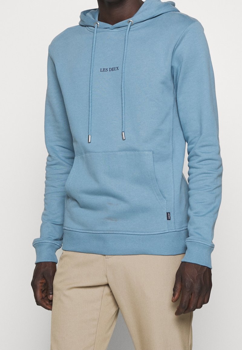 Homme portant un sweat à capuche bleu clair avec le texte « LES DEUX » sur la poitrine et un pantalon beige, debout devant un fond blanc uni.