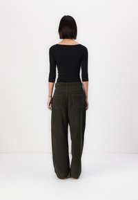 Zwarte langwerpige top in combinatie met donkergroene, wijde broek met twee achterzakken. De stoffen lijken zacht en gestructureerd.