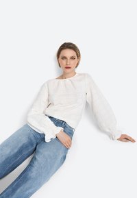 Haut à manches longues blanc avec un motif texturé et des poignets froncés, associé à un jean taille haute bleu clair. Design et silhouette minimalistes.