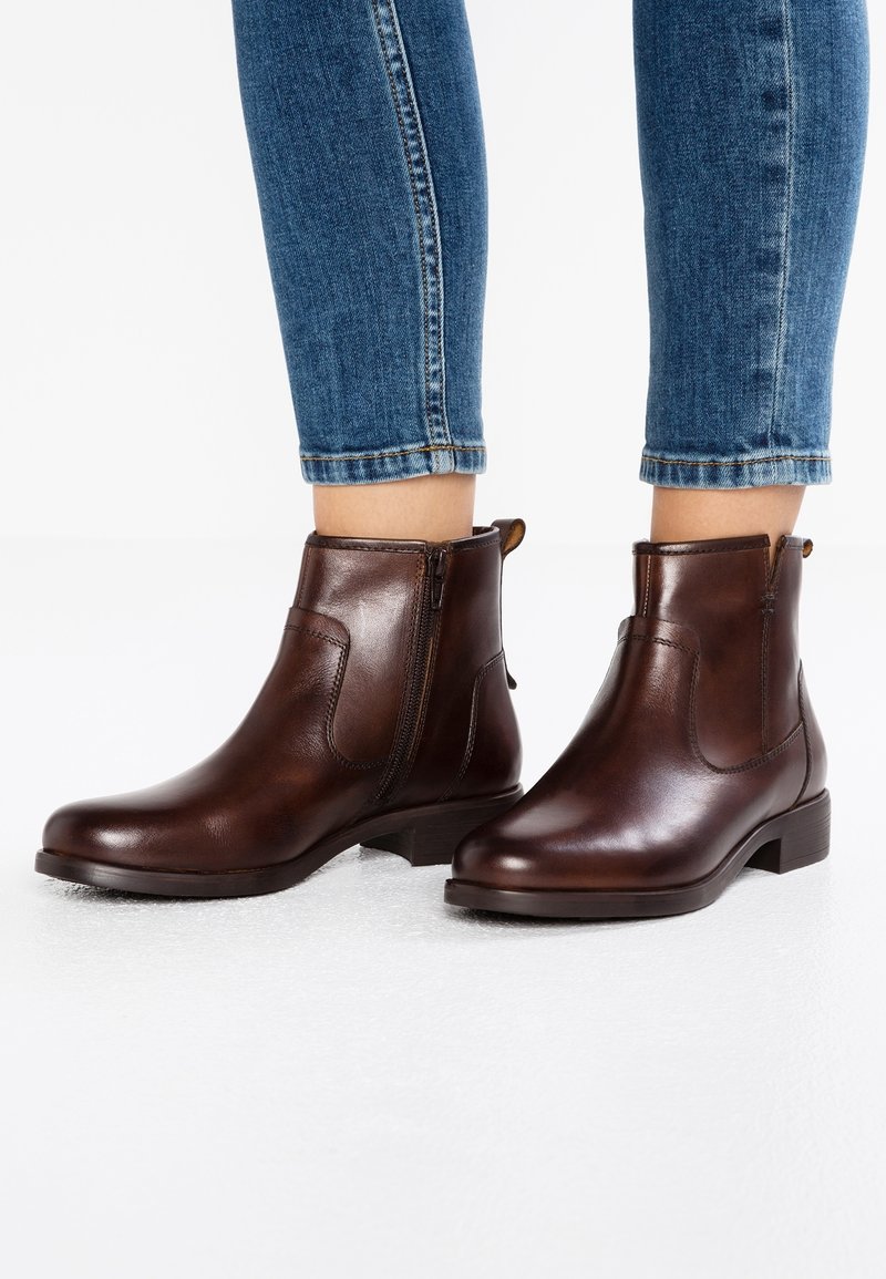 Bottines en cuir brun foncé avec des zips sur les côtés, portées avec un jean en denim bleu, se tenant sur un sol blanc.