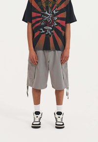 T-shirt grafica nera con un design rosso e giallo, abbinata a pantaloni cargo grigi chiari con tasche laterali e orli con cordino. Sneakers alte.